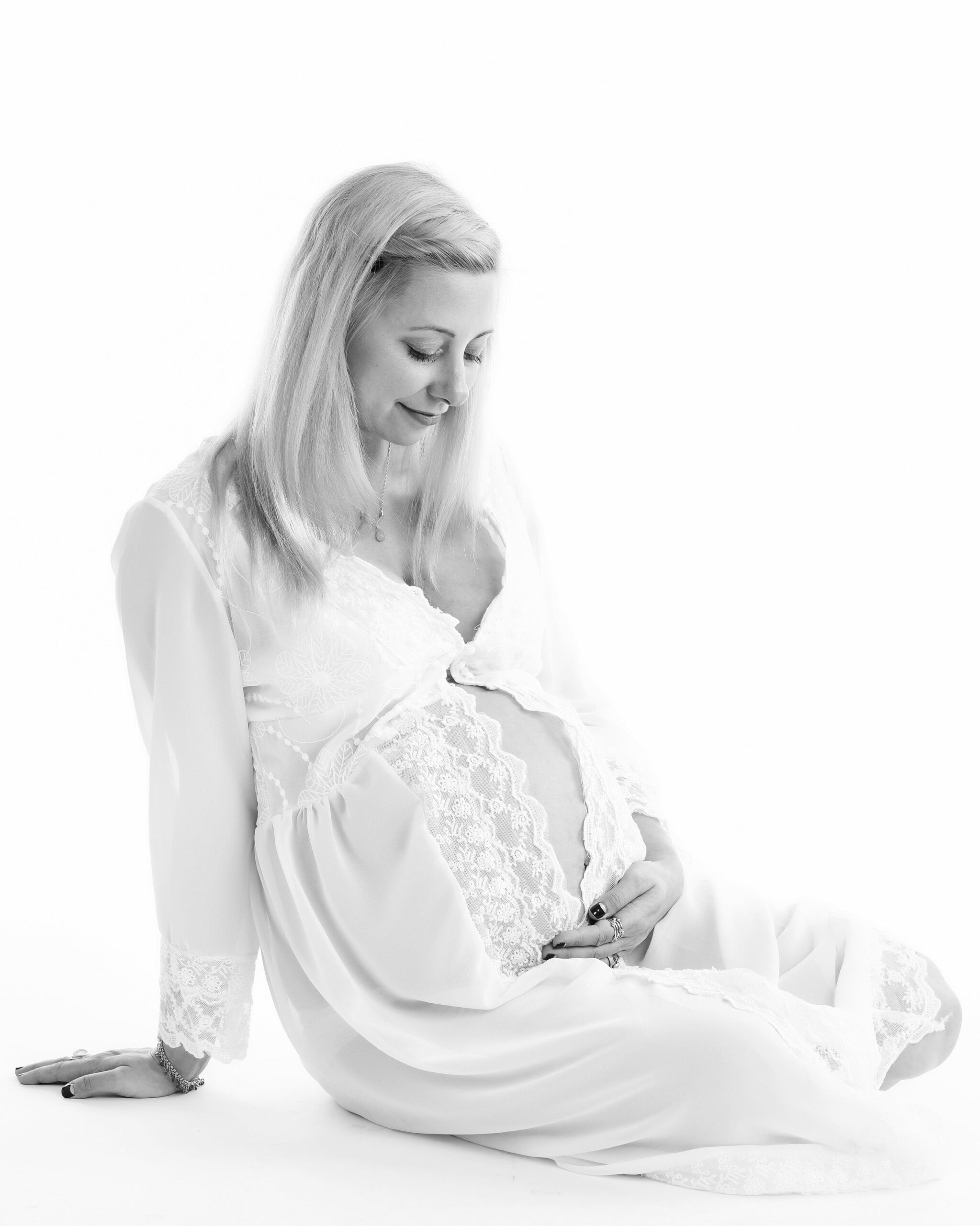 A pregnant woman in a delicate white gown sits on the floor, gently holding her belly and looking down with a peaceful smile, captured in a high-key lighting style. Toronto pregnancy portraits

