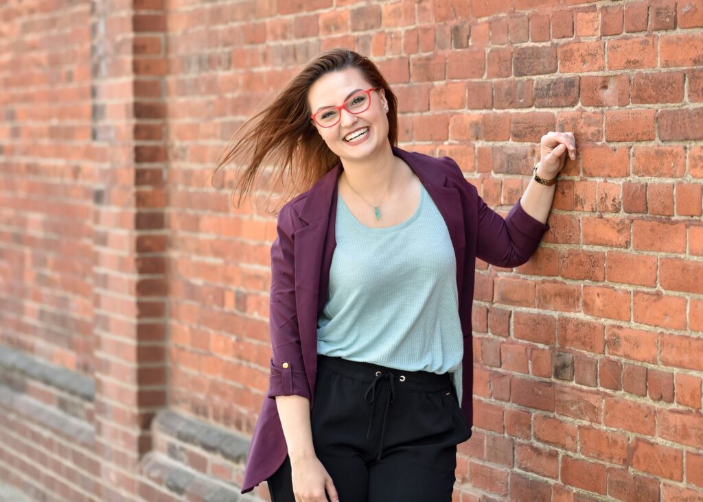 Woman with red glasses and a purple blazer smiling against a brick wall - how to take personal branding photos for Instagram.