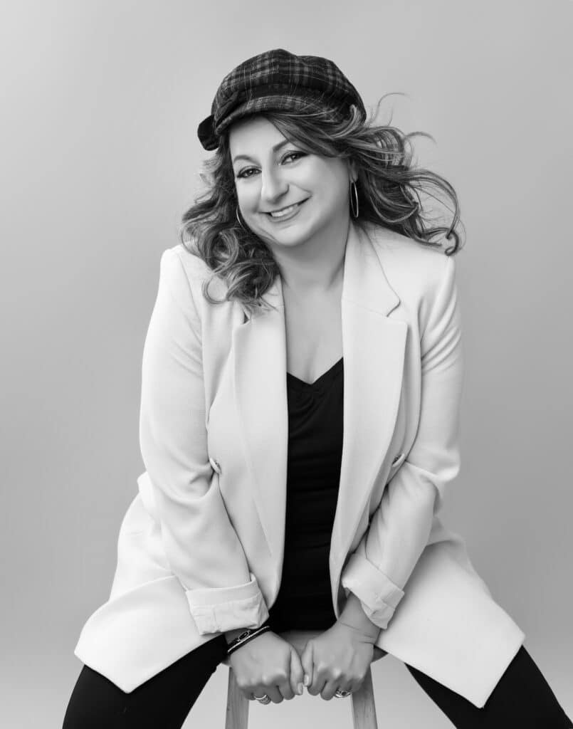 Black and white portrait of a smiling woman in a plaid cap and white blazer, seated editorial pose — Ellahrika Photography, Milton, Ontario
